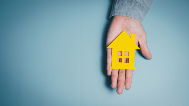 Single hand holding yellow house cutout, home security awareness, emotional balance, financial planning stability, supportive living insight, community safety care concept