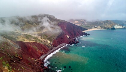 A dramatic coastal view of a rugged, red-colored cliff meeting the ocean under a cloudy sky