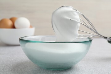 Beating egg whites in bowl and whisk on grey textured table, closeup