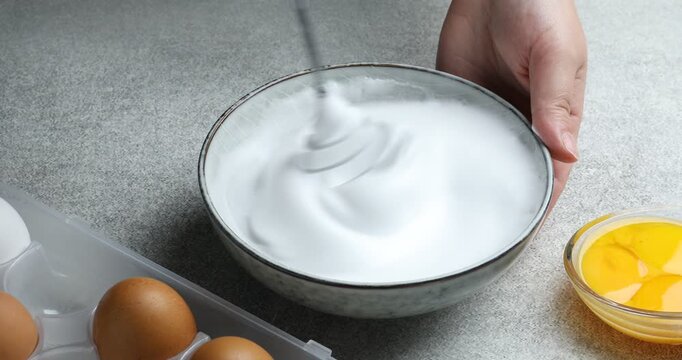 Woman whisking egg whites in bowl at grey textured table, closeup