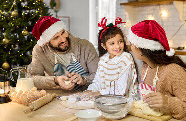 Christmas happy family baking cookies in kitchen. Parents help child daughter cut dough beside the festive tree, shaping a warm holiday mood at home. New Year celebration and traditions.