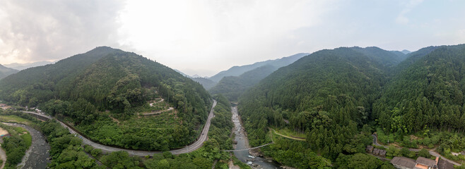 Panoramic view of a winding road through a lush, green valley flanked by mountains. A river flows alongside the road, reflecting the cloudy sky. A small building is visible on the right.