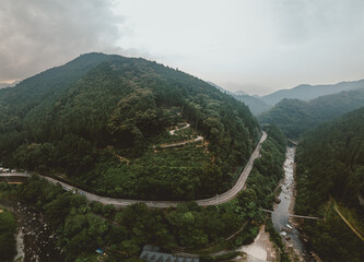 Aerial view of a winding road through a lush, mountainous landscape. A river flows alongside, framed by dense forests under a cloudy sky. Dramatic and serene.