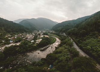 Aerial view of a winding river valley with lush green forests and a small town nestled along the banks. A road follows the river's course through the mountains. Overcast sky adds a moody atmosphere.