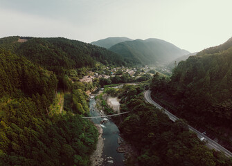Aerial view of a mountainous valley with a river, road, and small town. Lush green forests dominate the landscape under a cloudy sky. Serene and picturesque.