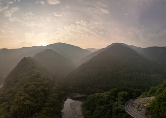 A stunning aerial view of a mountain range at sunrise, with a winding road cutting through a lush valley and a river flowing through it. The sky is filled with dramatic clouds.