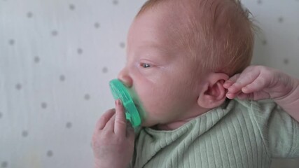 baby chewing green pacifier attentively, newborn sucking on bright green teether calmly, young infant with green pacifier as caregiver gently adjusts ear in cozy nursery environment