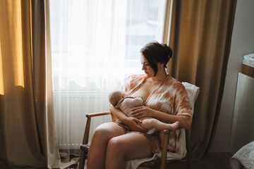 A mother sits in a wooden chair, gently breastfeeding her baby in a warm and softly lit room. Sunlight filters through the curtains, creating a peaceful atmosphere.
