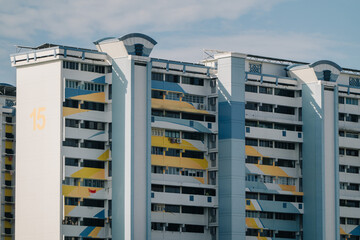 A colorful HDB block in Singapore with a distinctive geometric design. The building features a mix of white, yellow, and blue, with a prominent "15" on one side. The sky is blue with some clouds.