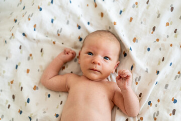 A newborn baby, lying on a soft, colorful blanket, gazes curiously at the viewer with wide eyes. The warm atmosphere highlights the baby's gentle features and innocence.