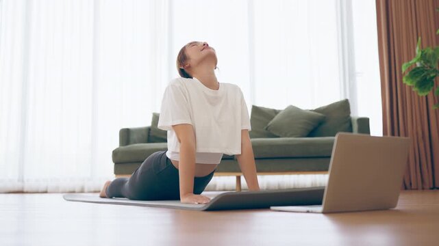 Young Asian woman stretching yoga workout on exercise mat while online training class with computer laptop in living room