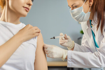 Close up shot of doctor in protective mask and gloves giving vaccination to young woman. Female medical worker with syringe administering injection during health appointment. Immunization concept.