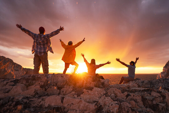 Group of happy diverse tourists are standing and sitting with open arms at sunset lake shore.