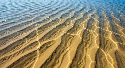 Fototapeta premium Close-up of golden sand with gentle wave patterns in shallow water, creating abstract textures