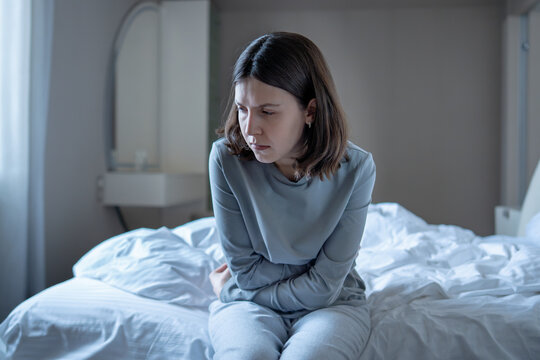 Depressed young woman seated on unmade bed arms crossed over stomach. Anxious stressed female alone in bedroom deep in bad thoughts struggling with emotional pain, uncertainty, mental health issues