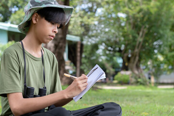 The 15-year-old Thai boy is captured using his binoculars to scan the distance while sitting on the grass, embodying the action of nature exploration and surveying his park surroundings.