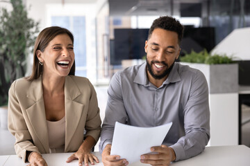 Two cheerful multiethnic coworkers discussing paper document at workplace