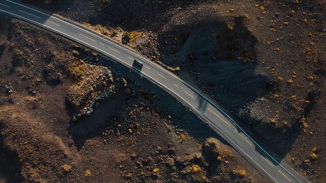 Winding road through rugged volcanic terrain with a single black vehicle. Extreme travel, adventure, drone shot, and harsh environment.