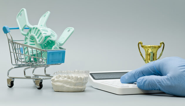 Mini shopping cart filled with dental impression trays beside a jaw model, calculator, and trophy, symbolizing cost, quality, and dental excellence.