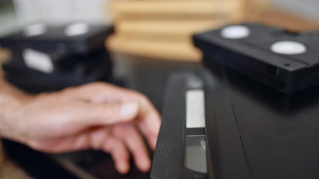 VHS cassette on VCR surface. A videotape rests partly inserted into a VCR with a blurred television behind it.