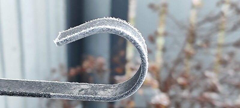A close-up of a frosted metal hook with a blurred background of winter foliage. The hook is curved and covered in ice crystals, indicating cold weather. - Powered by Adobe