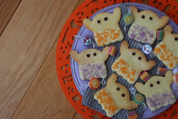 Top view of Halloweeen shortcrust cookies in shape of a ghost with white chocolate glaze and  sprinkles on a lilac plate on orange festive underplate  on wooden table 