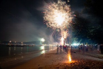 Long exposure shot capturing large white and gold fireworks exploding above a sandy beach with sparks rising from the ground and dark silhouettes of people watching
