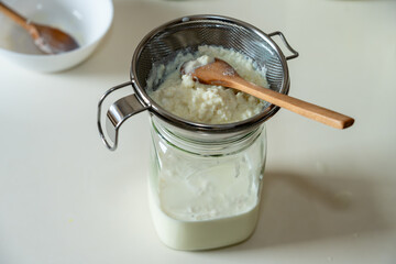 Overhead shot of white milk kefir grains in a strainer being gently stirred with a wooden spoon above a glass jar containing the freshly prepared fermented drink