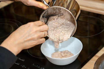 Close up of a persons hands serving a thick mixture of cooked porridge or oatmeal from a steel pot...