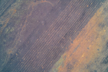 Top-down aerial photo of tilled agricultural soil with strong diagonal linear patterns. Muted cold purple gray tones contrast with warm rusty patches. Concept for agriculture, climate.