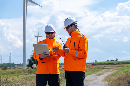 Two workers in bright jackets analyze data near wind turbines on a sunny day in a rural area