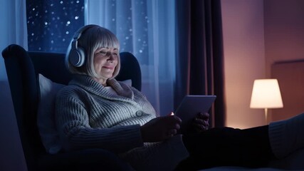 Senior woman using tablet and headset. Relax at home in evening light. Smile shows contentment with technology. Cozy sweater and soft lamp create warm calm. Window reveals gentle snow outside now.