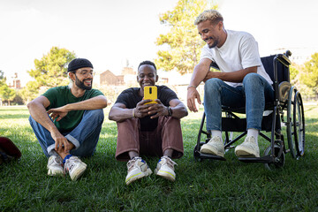 African American man in a wheelchair and friends sitting on the grass looking at mobile phone in a...