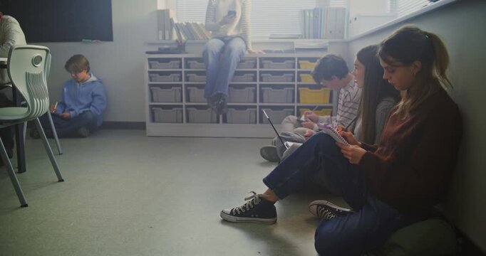 Several Students Sitting on Floor, Actively Writing in Notebooks and Sharing Laptop. Concept Flexible Learning Spaces, Peer To Peer Collaboration, and Comfortable Educational Environment. Static Shot.