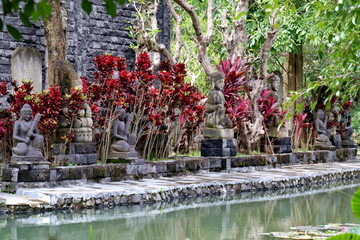 Stone statues sit peacefully along a pond surrounded by lush tropical plants in a serene Balinese...