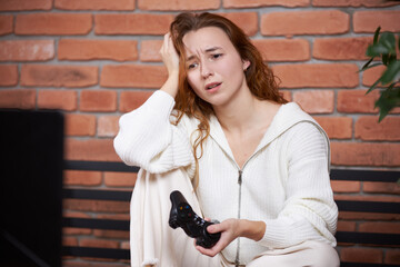 A young beautiful girl sits on the sofa holding a gamepad in her hands and playing a computer game. The player is sad because of losing the game