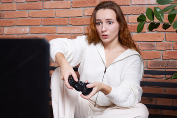 A young beautiful girl sits on the sofa holding a gamepad in her hands and playing a computer game.