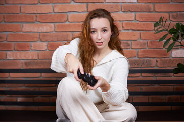 A young beautiful girl sits on the sofa holding a gamepad in her hands and playing a computer game.
