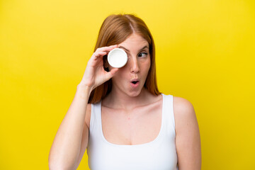 Young redhead woman isolated on yellow background with moisturizer and surprised