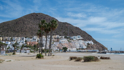 San Jose Beach in Almeria, Spain. A sandy shore with palm trees, a marina, and the white hillside village of San Jose against a rocky mountain under a blue sky