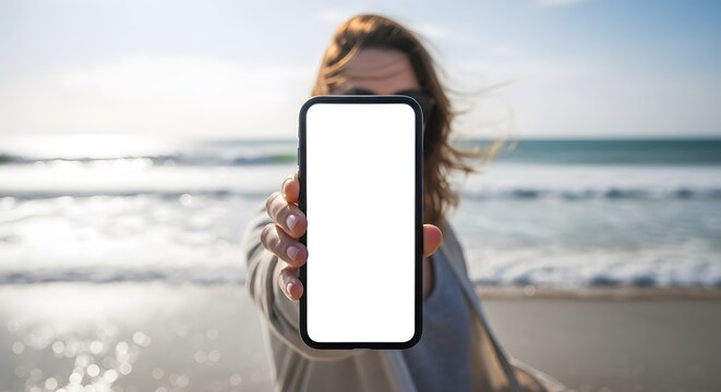 A woman holds up a smartphone with a blank screen in front of a beach with waves and a clear sky. - Powered by Adobe