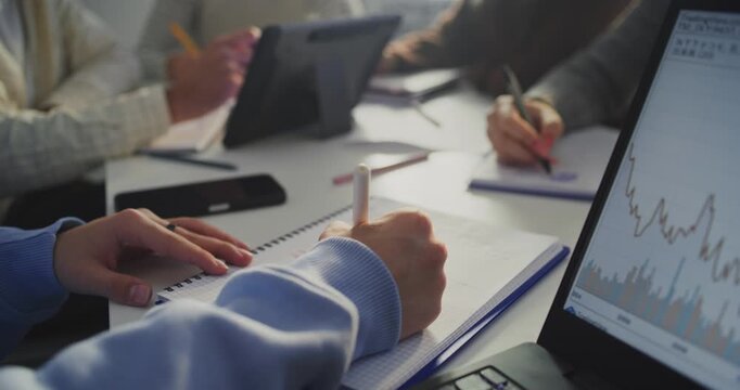 Close Up Hands of Several Students Intently Working at Shared Table. Student Writing in Notebook Next to Laptop Screen Displaying Analytical Graphs. Concept of Data Analysis, Digital Learning.