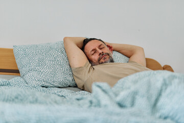 Middle-aged man relaxing and sleeping in bed on a cozy morning in a light-filled bedroom