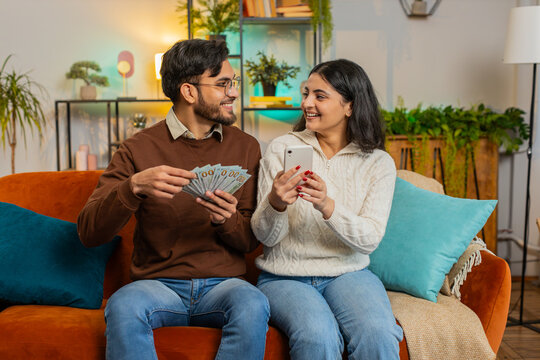 Happy Indian couple holding money cash and smartphone planning discussing investment together sitting on sofa in living room at home. Successful smiling diverse family winning lottery jackpot game