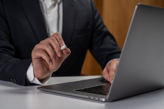 Businessman using wireless earbud while working on laptop at desk in office environment, focusing on technology and modern work concept.