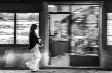 Black and white street photography woman walking past city shop window at night motion blur candid urban scene modern lifestyle smartphone fashion travel photo art european night city lights style