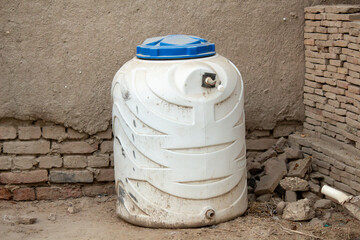 Large plastic water storage tank placed beside an old brick wall in a rural outdoor environment