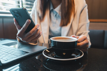 Professional woman multitasking with smartphone and laptop, enjoying coffee in a stylish cafe environment.