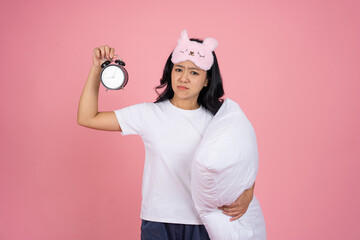 A tired and sleepy Asian woman holds an alarm clock and a pillow, standing isolated on a pink background.