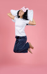 Young Asian woman in pajamas and an eye mask is jumping high with a pillow on a pink studio floor.